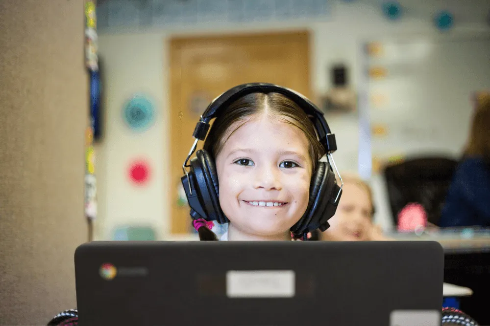 A smiling primary school student wearing headphones, working on a laptop in class